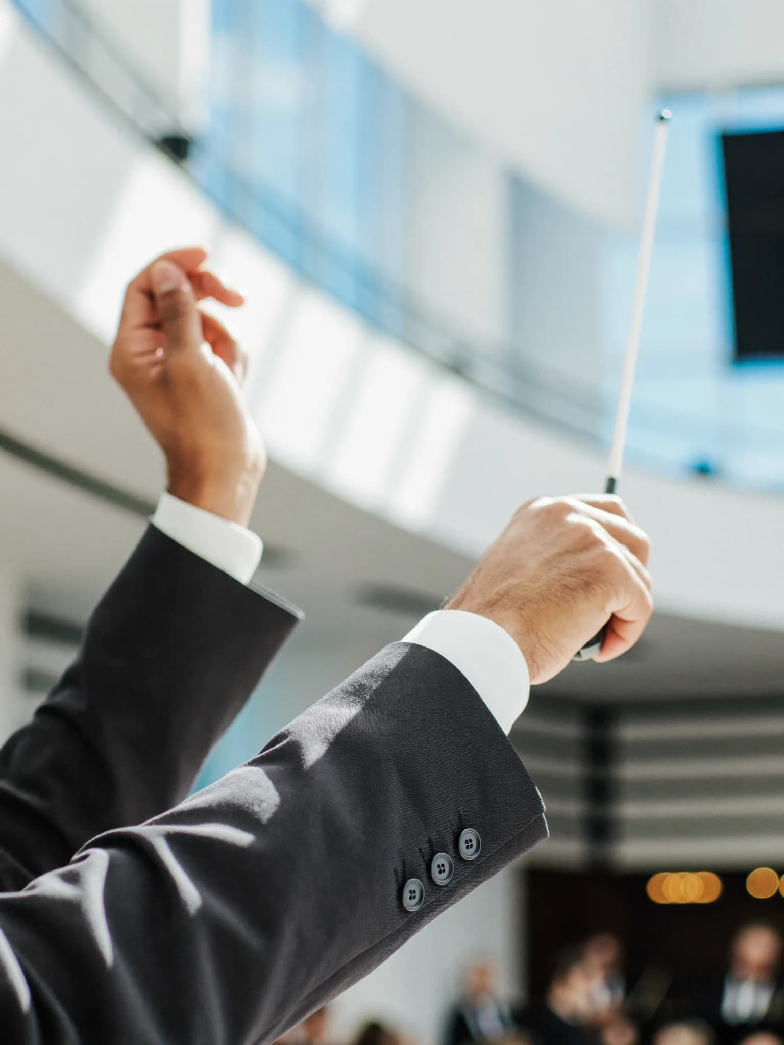 A conductor's raised hands holding a baton against a bright, softly blurred concert hall